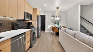 A modern kitchen with wooden cabinets, stainless steel appliances, and a dining area with a round mirror and a sofa. - Welcome to The Desert Rose Guesthouse – Where Desert Skies Meet Rooftop Vibes (Hurricane)