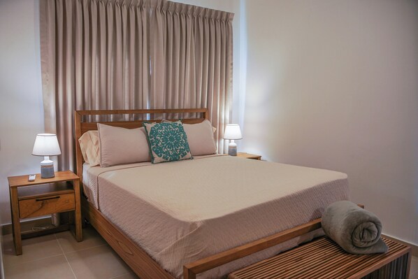 A neatly made bed with a wooden headboard, two bedside tables with lamps, and a rolled towel on a wooden bench.