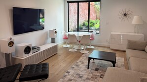 Una sala de estar moderna con una televisión de pantalla plana, parlantes, una mesa de centro, un sofá, una mesa de comedor con sillas y un ventanal con vista a un muro de ladrillo y vegetación.