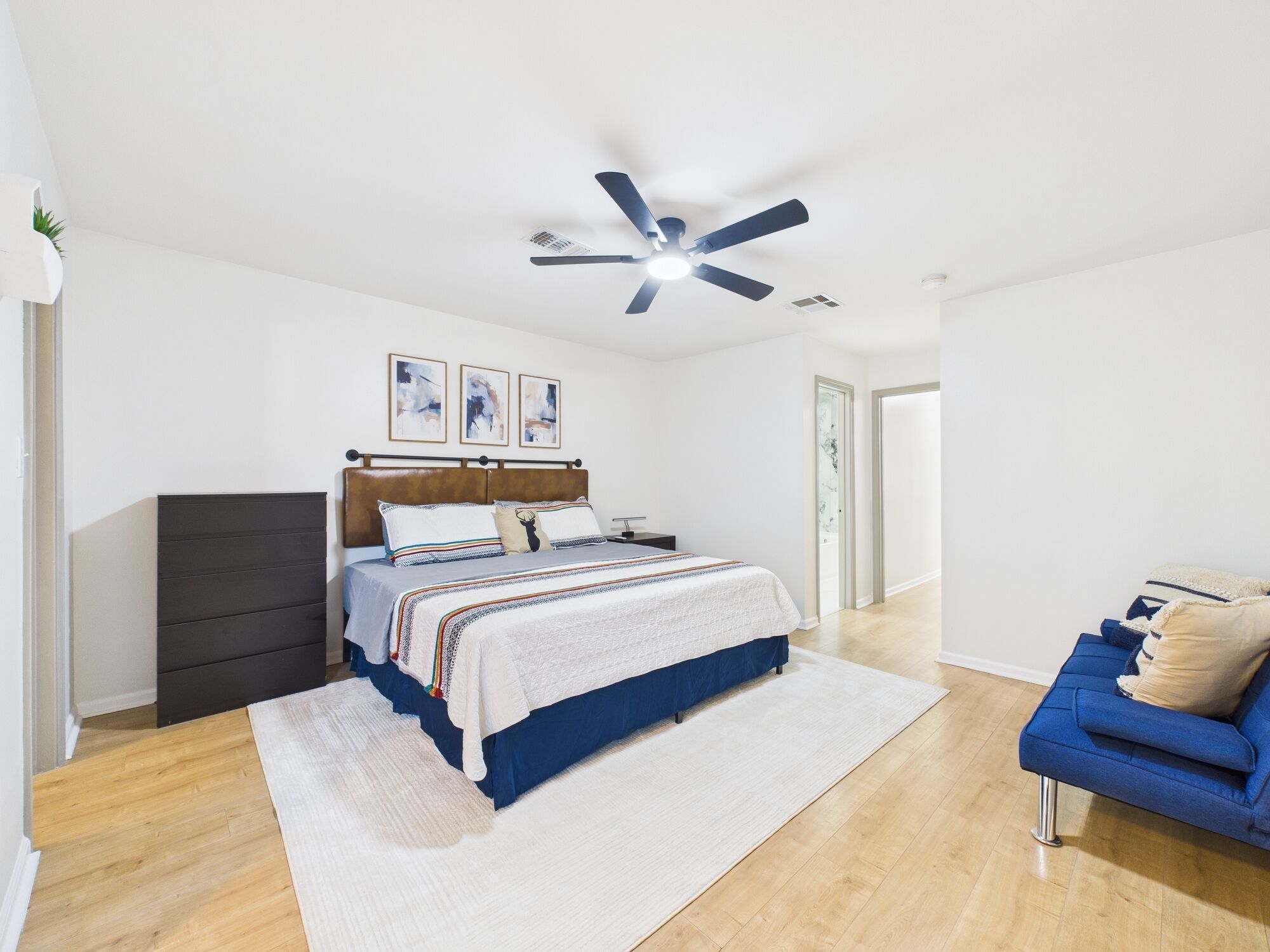 Una camera da letto con un letto, una ventola a soffitto, una cassettiera, un divano blu e un tappeto bianco.