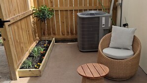 Eine Terrasse mit Holzlattenzaun, einem kleinen Blumenbeet, einem Korbstuhl mit Kissen und einem runden Tisch.