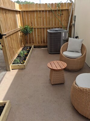 Eine Terrasse mit Holzlattenzaun, einem kleinen Blumenbeet, einem Korbstuhl mit Kissen und einem runden Tisch.