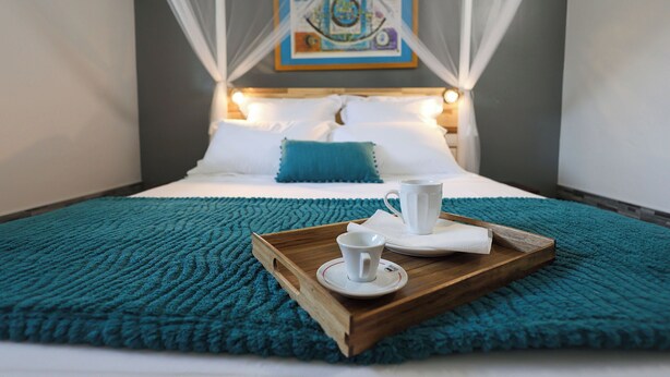 A neatly made bed with a blue throw blanket, a wooden tray with a white cup and saucer, and a painting above the bed. - Villa Écume Verte - 2 bedrooms, terraces, quiet & close to Sainte-Anne beaches (Sainte-Anne)