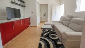 A modern living room with a sofa, a TV, and a kitchen area with red cabinets and a ceiling fan. - Villa Écume Verte - 2 bedrooms, terraces, quiet & close to Sainte-Anne beaches (Sainte-Anne)