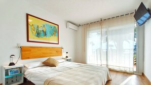 Ein Schlafzimmer mit einem hölzernen Kopfteil, einem Bett mit beiger Bettdecke, einem Nachttisch mit Lampe, einem Bilderrahmen und einem großen Fenster mit durchsichtigen Vorhängen.