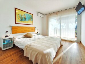 A bedroom with a wooden headboard, a bed with a beige bedspread, a nightstand with a lamp, a picture frame, and a large window with sheer curtains. - Beachfront apartment with swimming pool Miami Platja (Miami Platja)