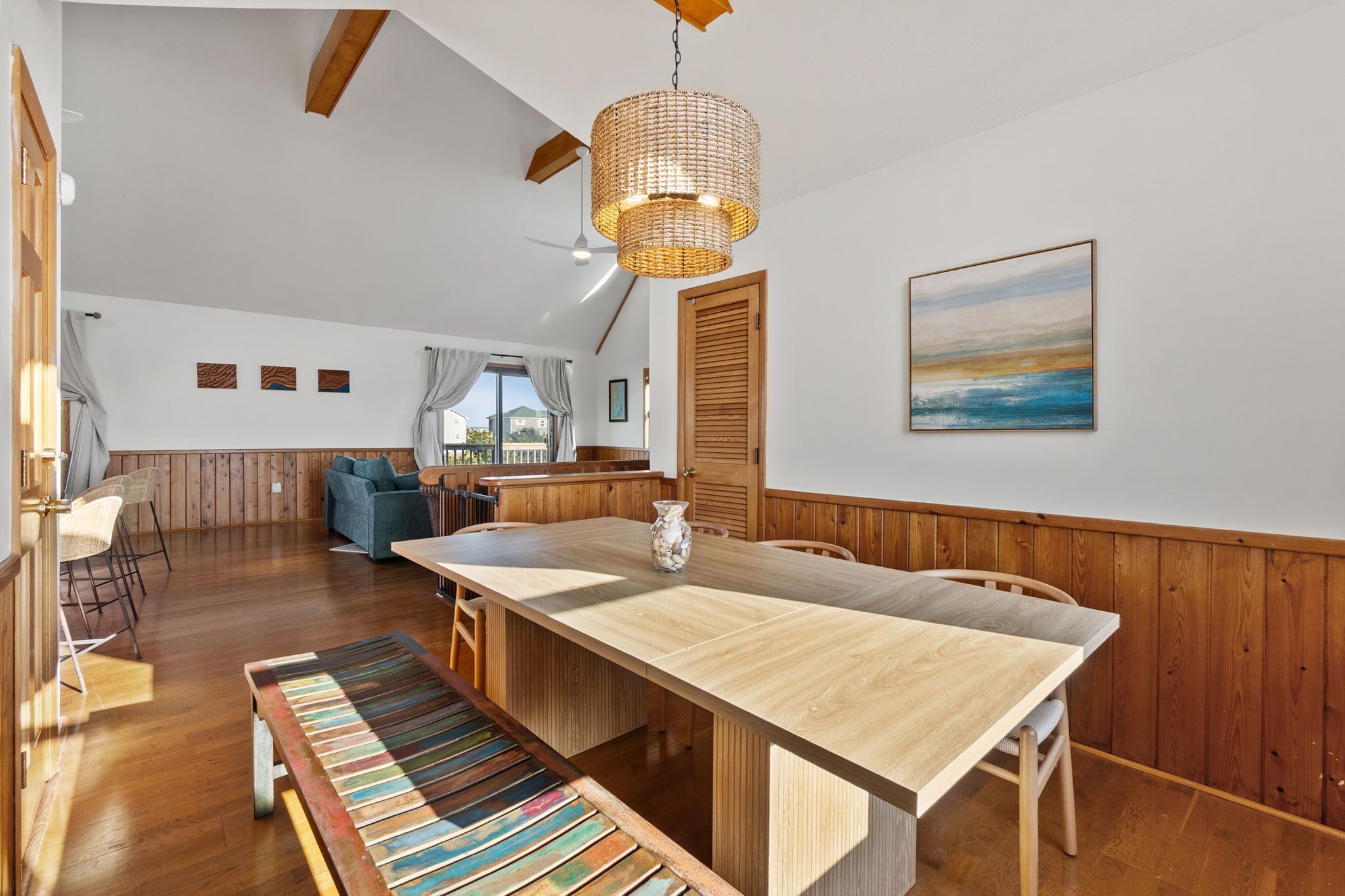 A spacious room with a wooden dining table, a bench, a hanging light fixture, and a painting on the wall.