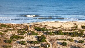 Een kustlandschap met een zandstrand, groene begroeiing en een helderblauwe lucht.