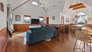 Een ruime woonkamer met een hoge plafond, houten balken, een grote bank, een eettafel met stoelen en uitzicht door een raam.
