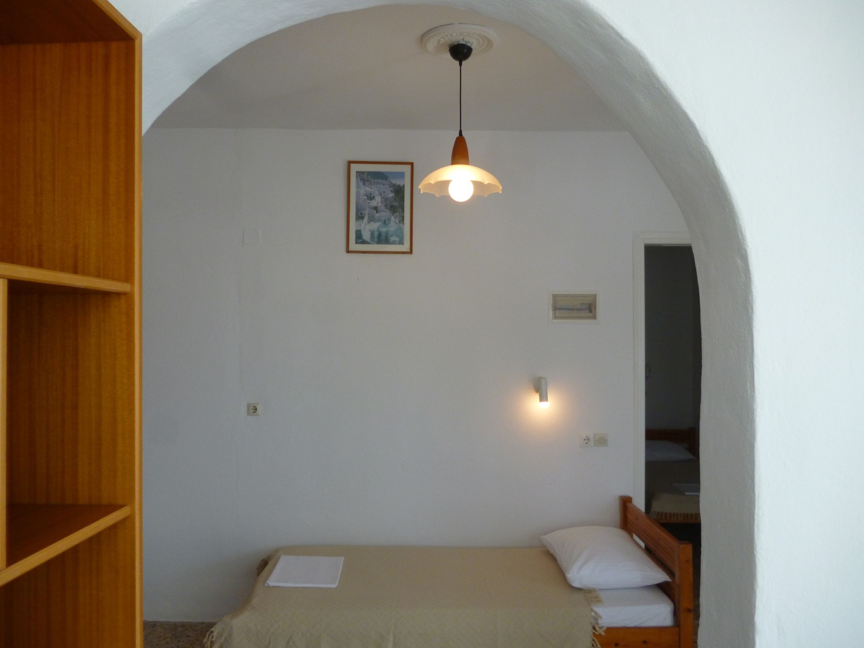 A room with a bed, a wooden shelf, a framed picture on the wall, and a hanging light fixture.