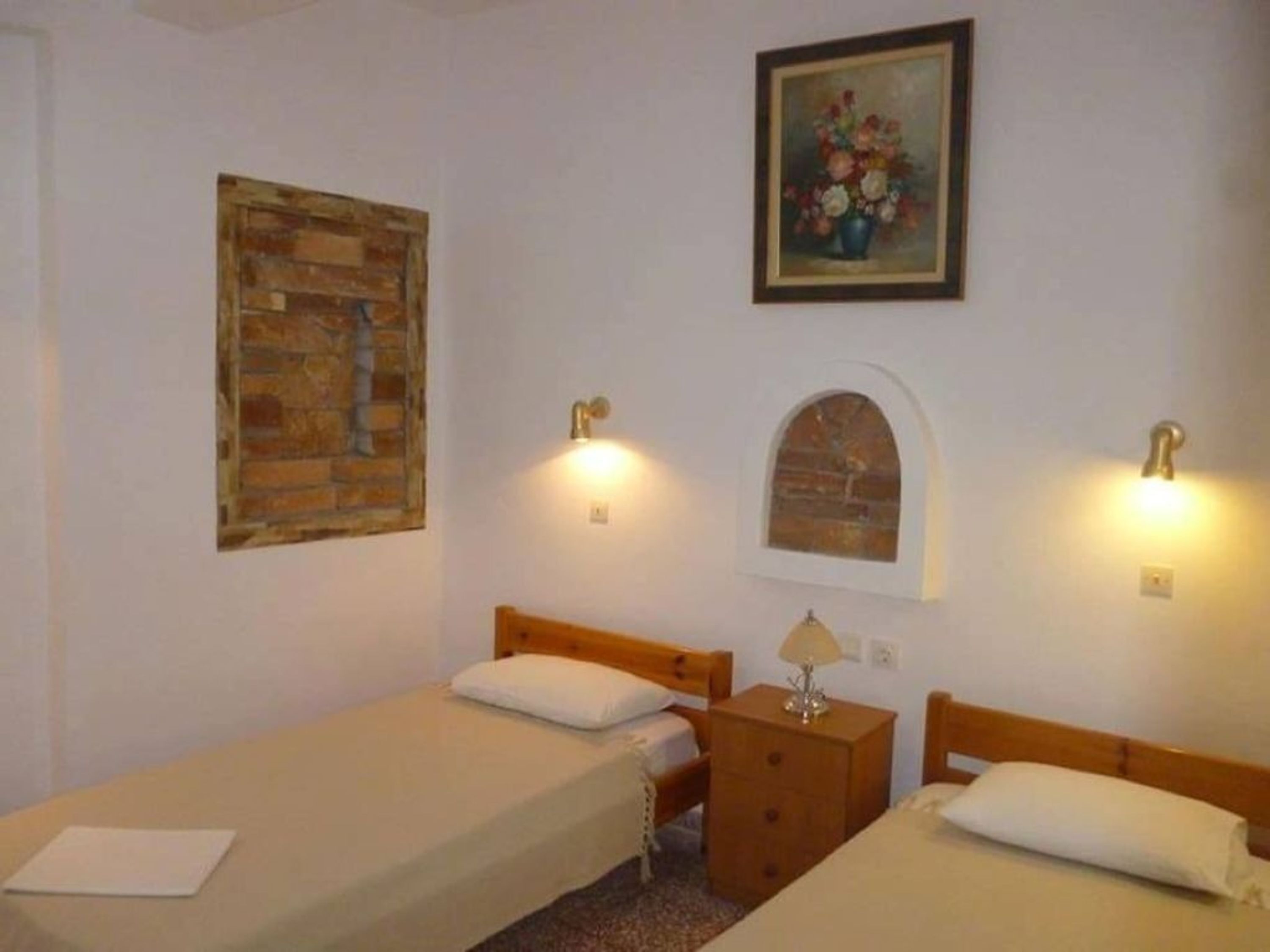 A room with two single beds, a wooden nightstand with a lamp, a brick-patterned wall, and a framed floral painting.