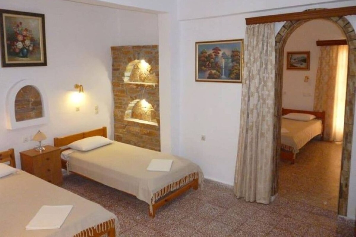 A room with two single beds, a wooden headboard, a stone wall with a shelf, a painting, and a doorway leading to another room.