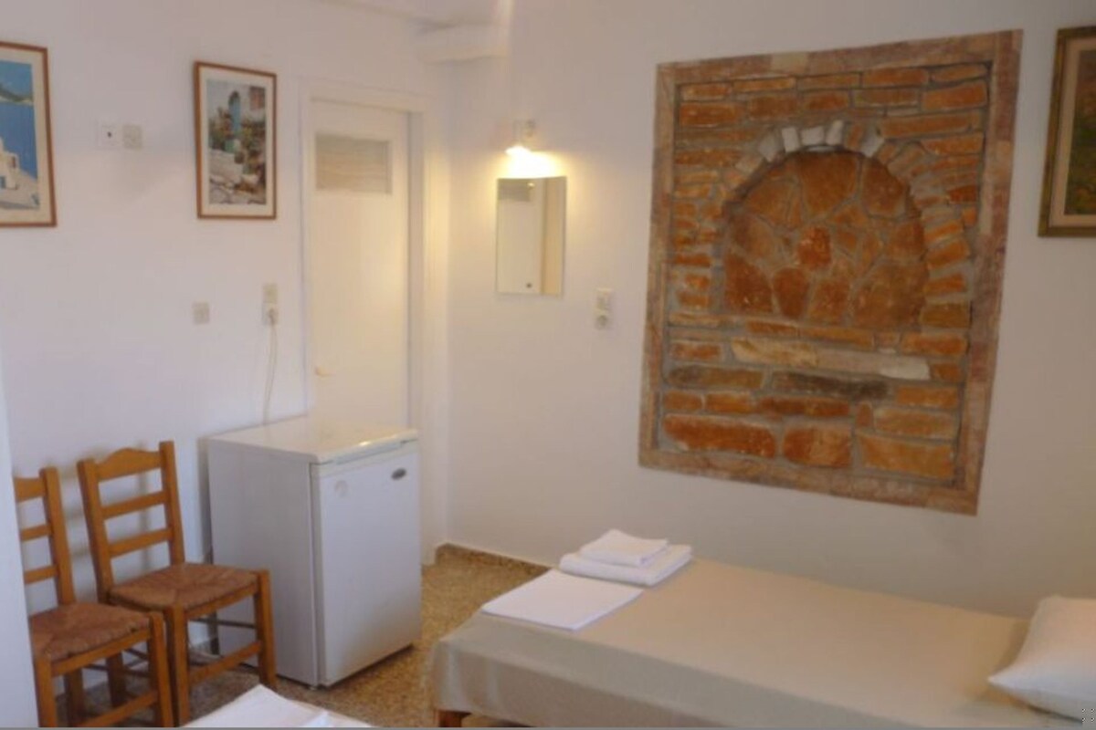 A small room with a bed, a desk, a chair, a refrigerator, and a wall-mounted fireplace.
