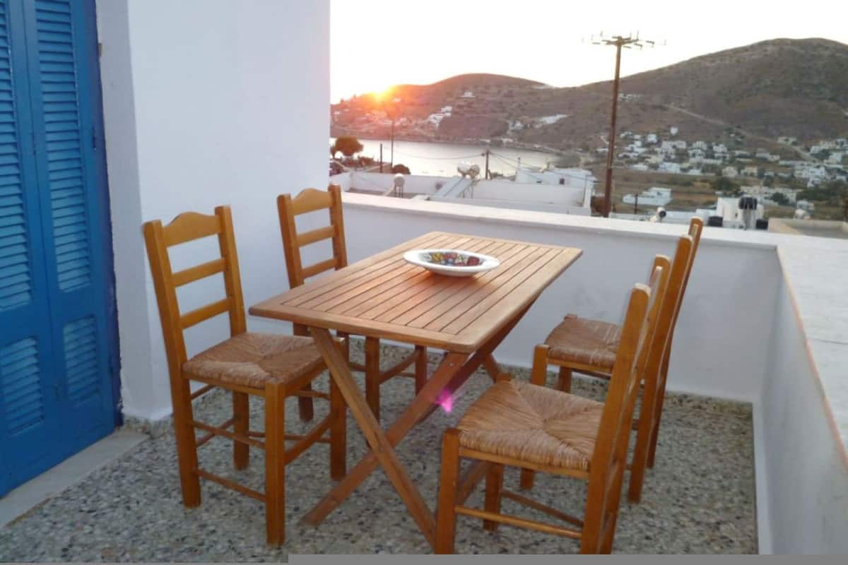 A wooden table with four chairs on a balcony with a view of a coastal town and hills.