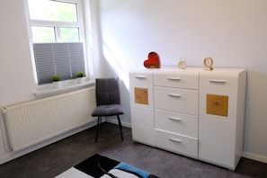 Ein Zimmer mit Heizkörper, einem Fenster mit Jalousien, einem grauen Sessel, einer weißen Kommode mit Schubladen und einem Teppich.