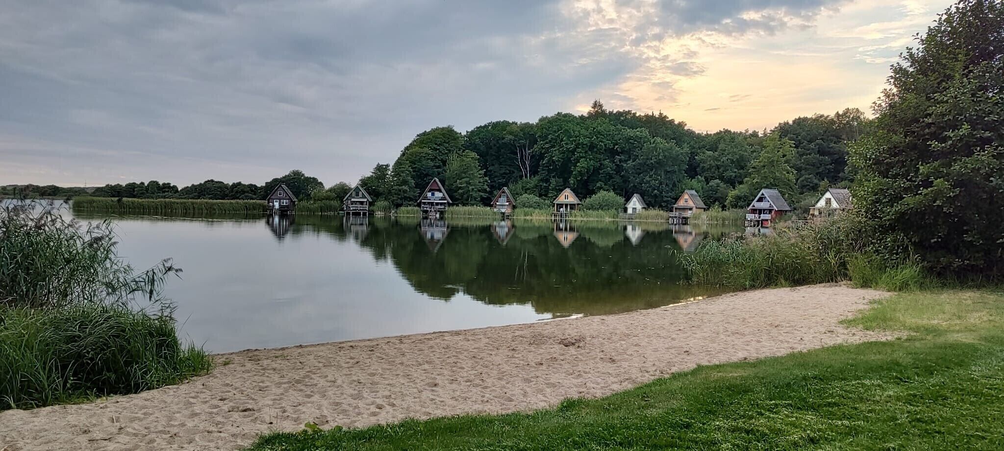 A tranquil lakeside view with several wooden structures on stilts, surrounded by lush greenery and a clear sky.