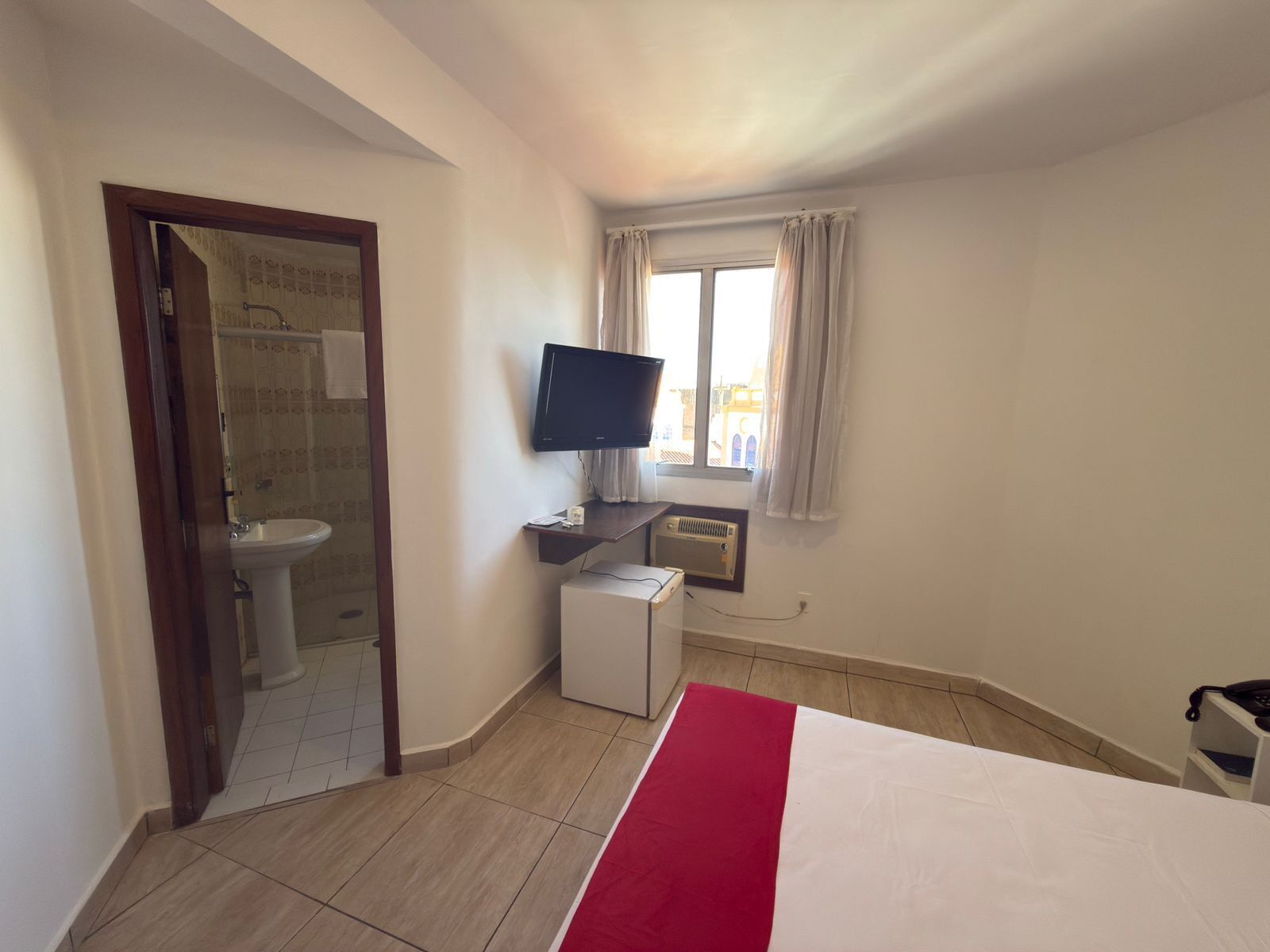 Habitación de hotel con cama, televisor, ventana con cortinas, lavamanos y aire acondicionado.