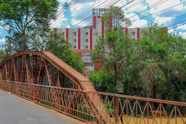 A bridge with a metal structure, a building with red and white vertical stripes, and greenery in the background.