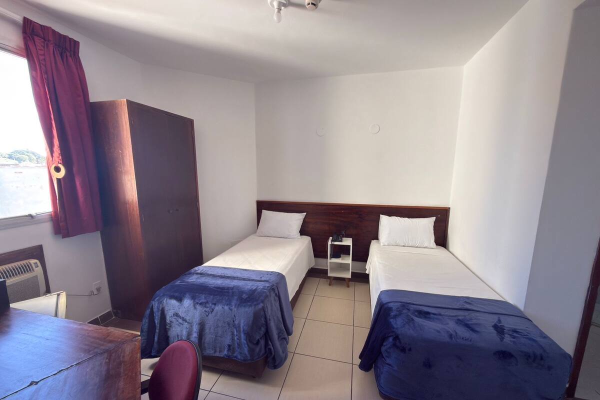 A hotel room with two beds, a desk, a chair, a wardrobe, and a window with curtains.
