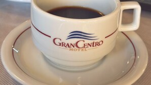 Een kop koffie met een logo op een schotel, een bord met gebak, en een laptop op de achtergrond.