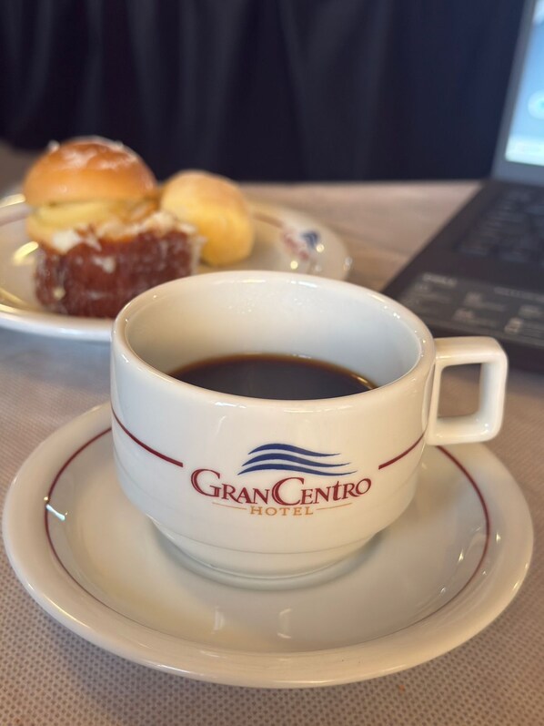 A cup of coffee with a logo on a saucer, a plate with a pastry, and a laptop in the background. - GRANCENTRO HOTEL (Mogi-Guacu)