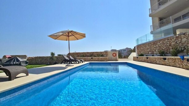 Una piscina con lettini e un ombrellone, un muro in pietra e un edificio moderno con balconi.