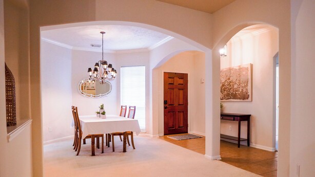A hallway with a dining area featuring a table and chairs, a wooden door, and a rug leading to a room with a painting. - Beautiful 3-bedroom house in The Woodlands (Spring)