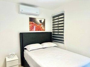 Ein Schlafzimmer mit einem Bett, einem Nachttisch mit einer Uhr, einer Klimaanlage, einem Fenster mit Jalousien und einem Bild an der Wand.