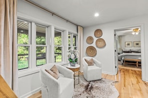 A room with large windows, a round table with a vase, two armchairs, a woven rug, and a bed with a headboard in the adjacent room. - Serene Lakefront Retreat w/ Private Hot Tub in Gated Community. (Blue Ridge)