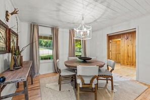 A dining area with a round wooden table, white upholstered chairs, a chandelier, and a rustic wall decor. - Serene Lakefront Retreat w/ Private Hot Tub in Gated Community. (Blue Ridge)