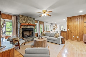 A living room with a stone fireplace, wooden walls, a ceiling fan, and a large window. - Serene Lakefront Retreat w/ Private Hot Tub in Gated Community. (Blue Ridge)