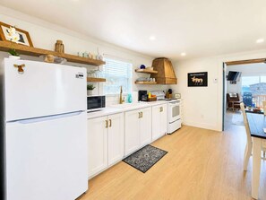 Une cuisine moderne avec des meubles blancs, un réfrigérateur, un four à micro-ondes et une cuisinière. Il y a une étagère en bois au-dessus de l’évier et une enseigne noire accrochée au mur.