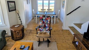 A living room with a wooden floor, a large window with a view, a chair with a colorful blanket, and a small table with a remote control. - Wild Cove Retreat & Nook (Twillingate)