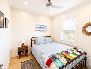 Une chambre à coucher comprenant un lit, une table de chevet, une fenêtre avec des stores, un ventilateur de plafond et un miroir fixé au mur.