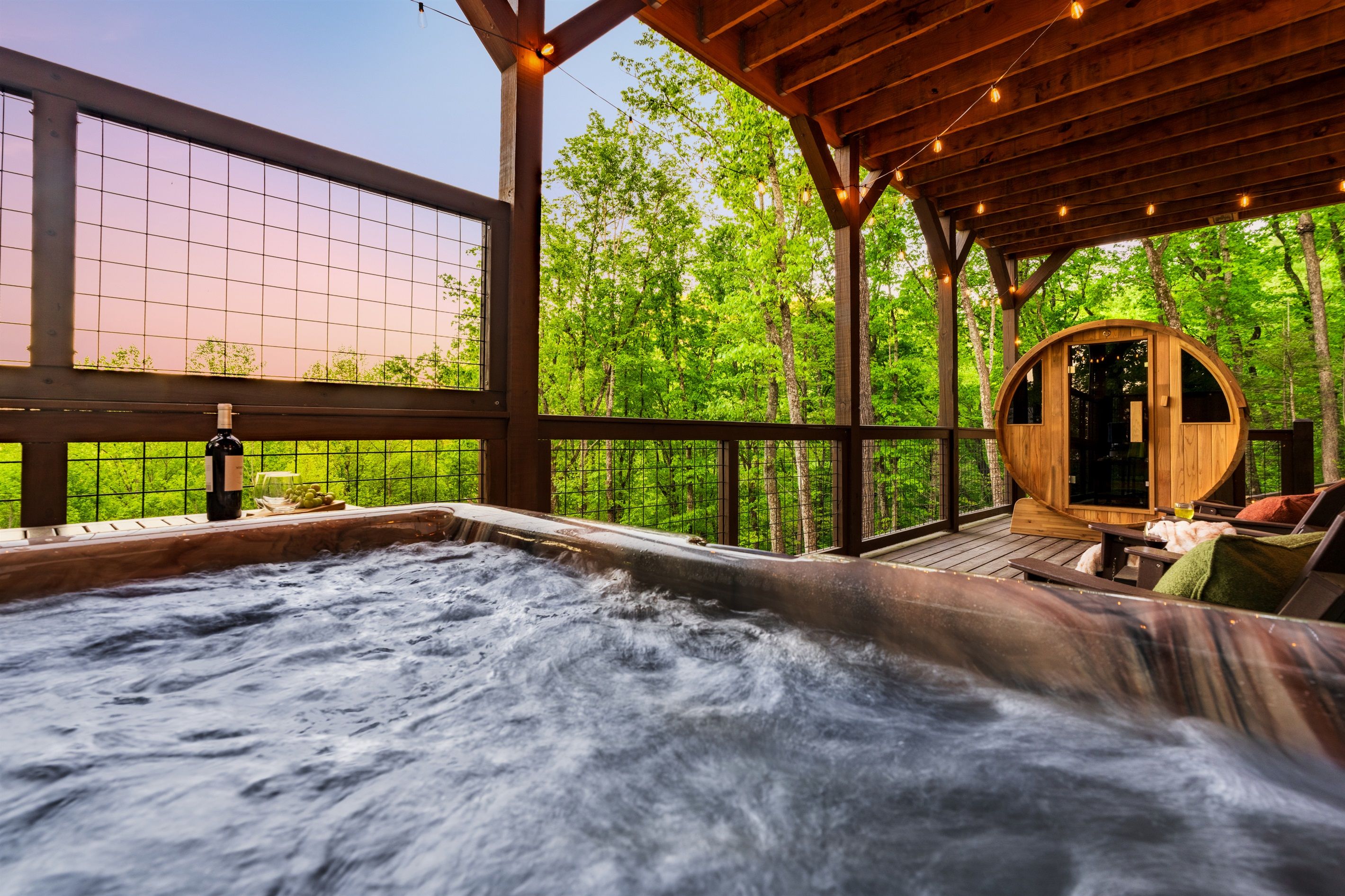 Un jacuzzi con terraza de madera, rodeado por una pérgola de madera con luces colgantes, con vistas a un bosque.