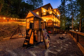 Eine Holzhütte mit Spielbereich, darunter eine Rutsche und eine Kletterwand, umgeben von Bäumen.