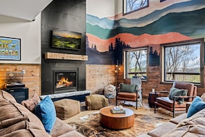 A cozy living room with a fireplace, comfortable seating, and a mural of mountains. - 5*Retreat w/Indoor Pool~FirePit~Arcade~Playground (Sevierville)