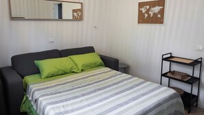 A bed with green pillows, a grey headboard, a striped bedspread, a small round table, a world map on the wall, and a black shelf unit.