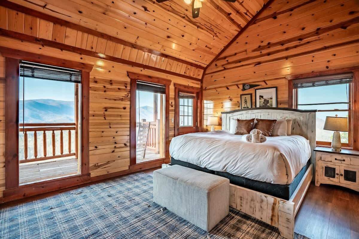 A wooden cabin bedroom with a large bed, a bench, and a view of the mountains.