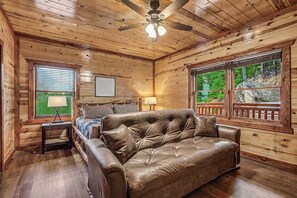 Ein gemütliches Blockhauss-Interieur mit einer Ledersofa, einem Bett, einem Deckenventilator und großen Fenstern, die einen Blick ins Freie bieten.