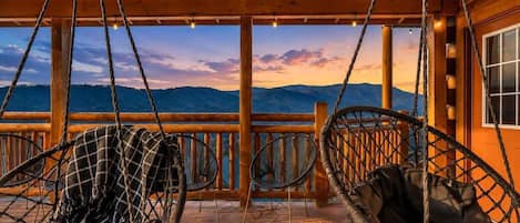 A wooden deck with a view of mountains, two wicker chairs, and a wooden railing.