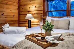 Ein gemütliches Zimmer mit Holzvertäfelung, einem Bett mit weißer Bettwäsche, einem Nachttisch mit Lampe, einer Ablage mit Keksen und einem Glas sowie einer Topfpflanze.