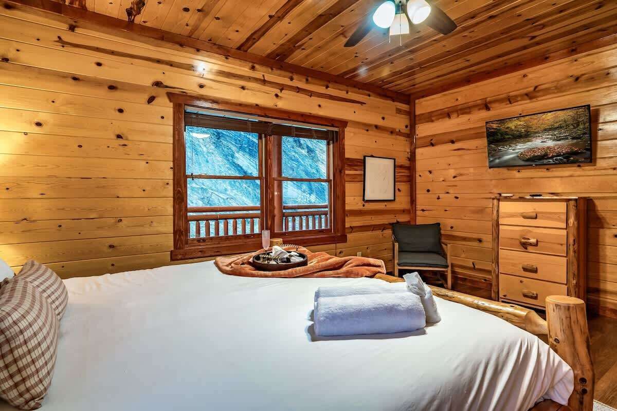 Une pièce de cabane en bois comprenant un lit, une chaise, une commode et une fenêtre donnant sur la forêt.