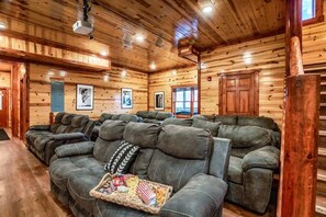 Ein Holzblockhaus-Interieur mit einem großen Ecksofa, Holzwänden und einem an der Decke befestigten Projektor.