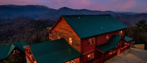 A log cabin with a green roof and a porch, surrounded by a mountainous landscape.