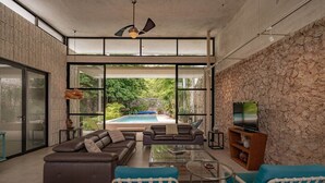 Ein großzügiger Wohnbereich mit einer großen Couch, zwei Sesseln, einem Couchtisch und einem Fernseher. Eine Glastür führt zu einem Poolbereich mit Blick ins Grüne.
