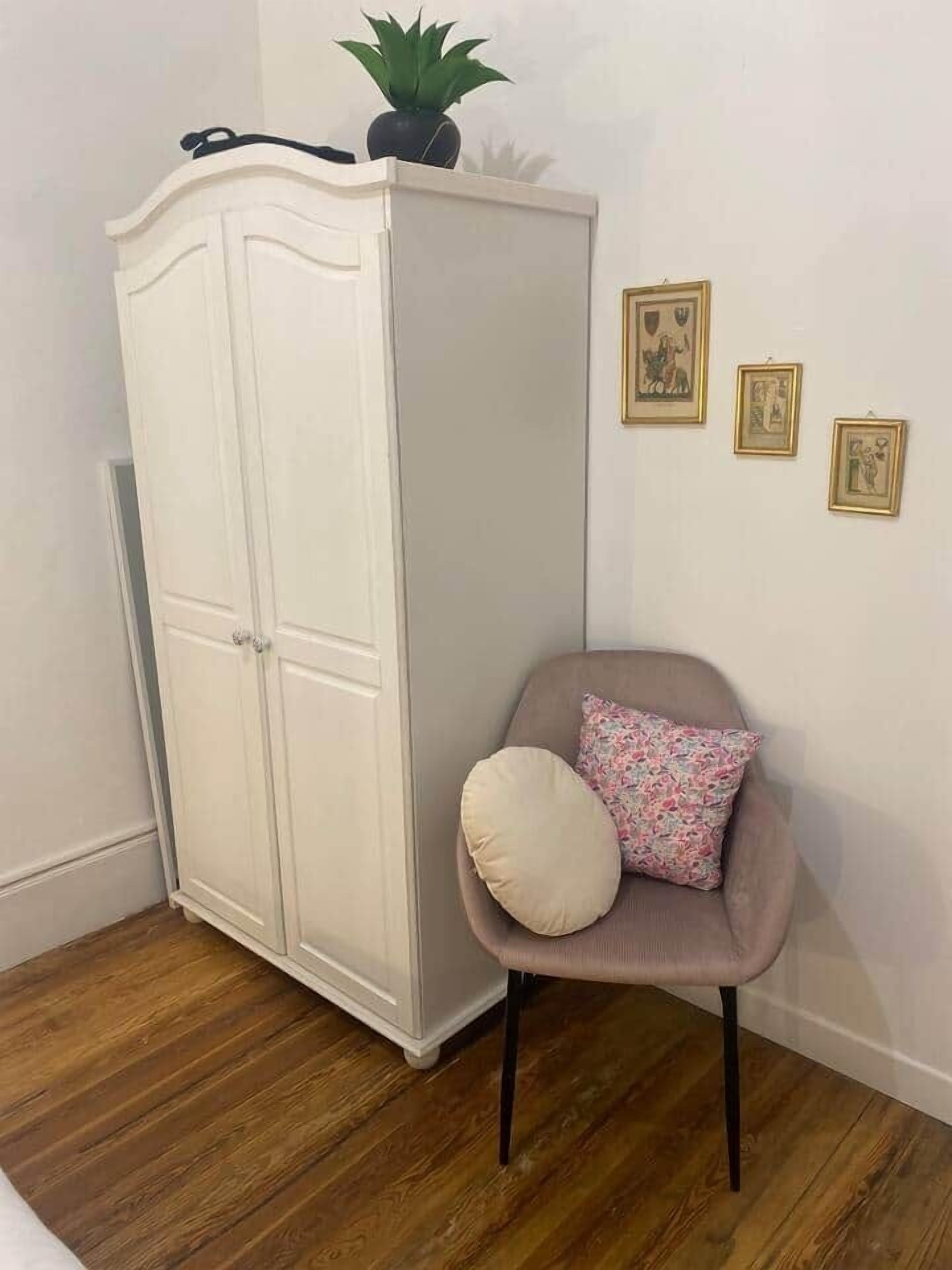 A room with a white wardrobe, a grey armchair with a floral pillow, and three framed pictures on the wall.