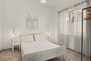 A bedroom with a bed, two bedside lamps, a wall-mounted macrame decoration, and a window with sheer curtains.