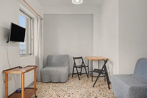 A small, sparsely furnished room with a television mounted on the wall, a single window, a small table, two chairs, and a sofa.