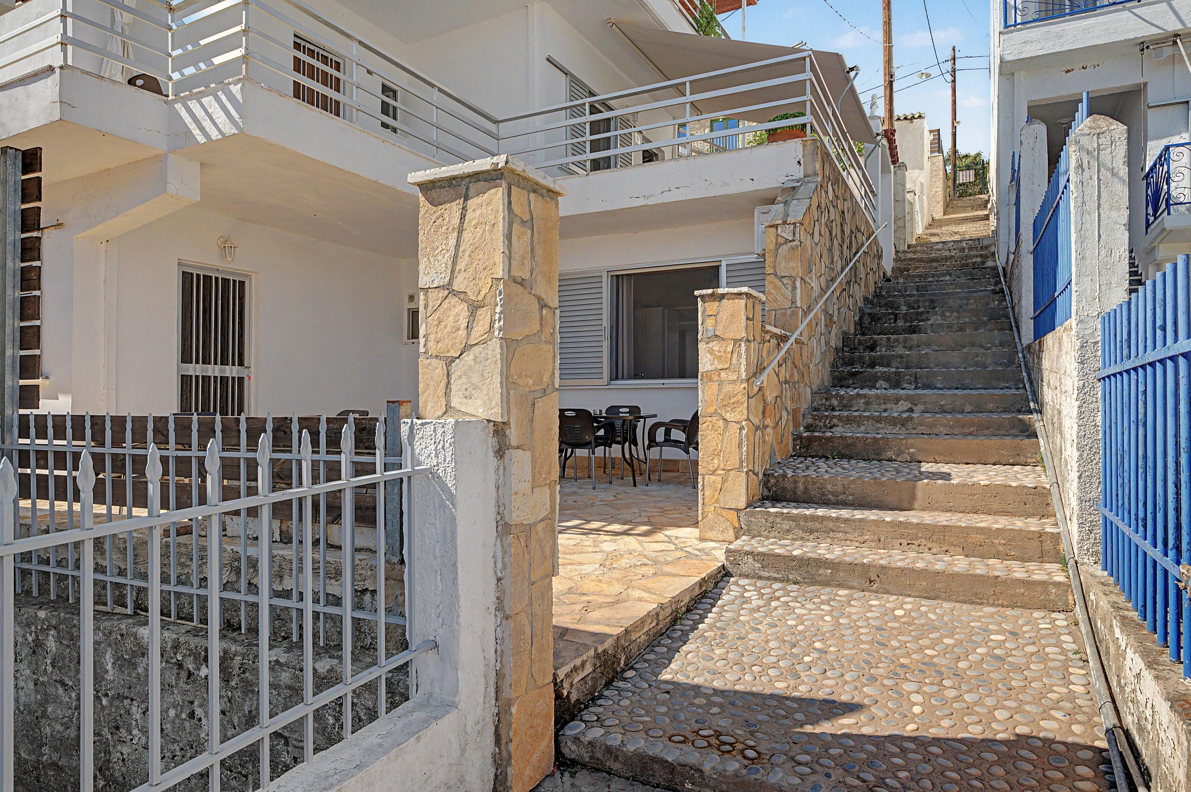 A residential building with a stone staircase leading to a balcony, a fenced patio area with outdoor furniture, and a white exterior.